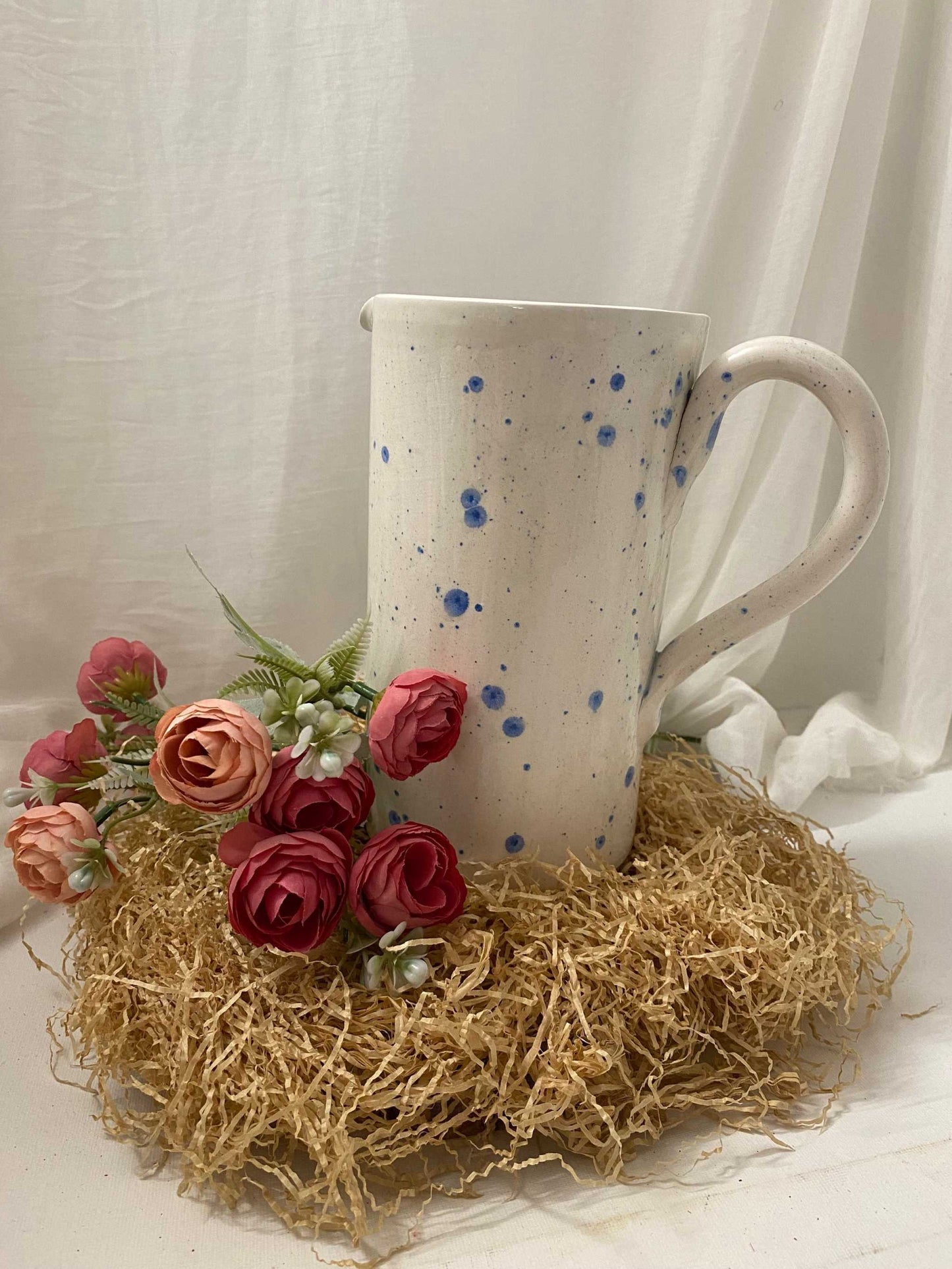 White ceramic pitcher with blue speckles from summer collection displayed with artificial flowers on straw mat