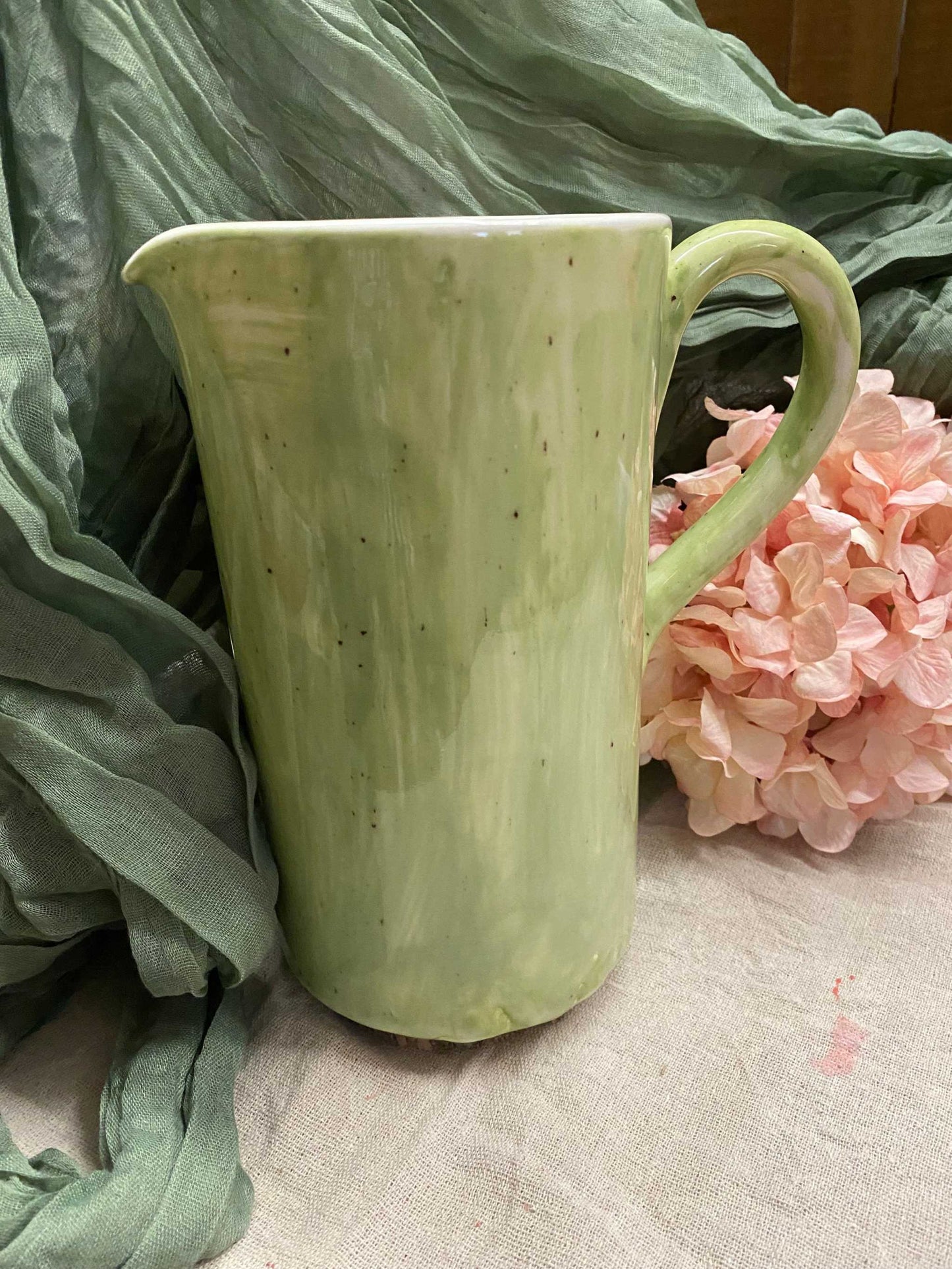 Light green ceramic pitcher with handle and spout placed near pink flowers and green fabric background
