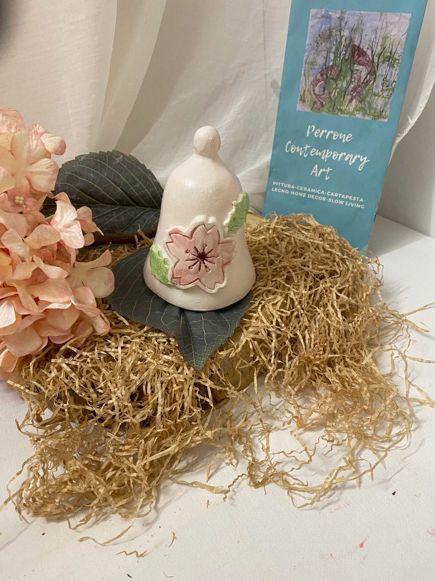 White ceramic bell with floral decoration from Campanella della Collezione Ciliegio placed on natural straw and dark leaves with pink flowers and a blue art flyer in background