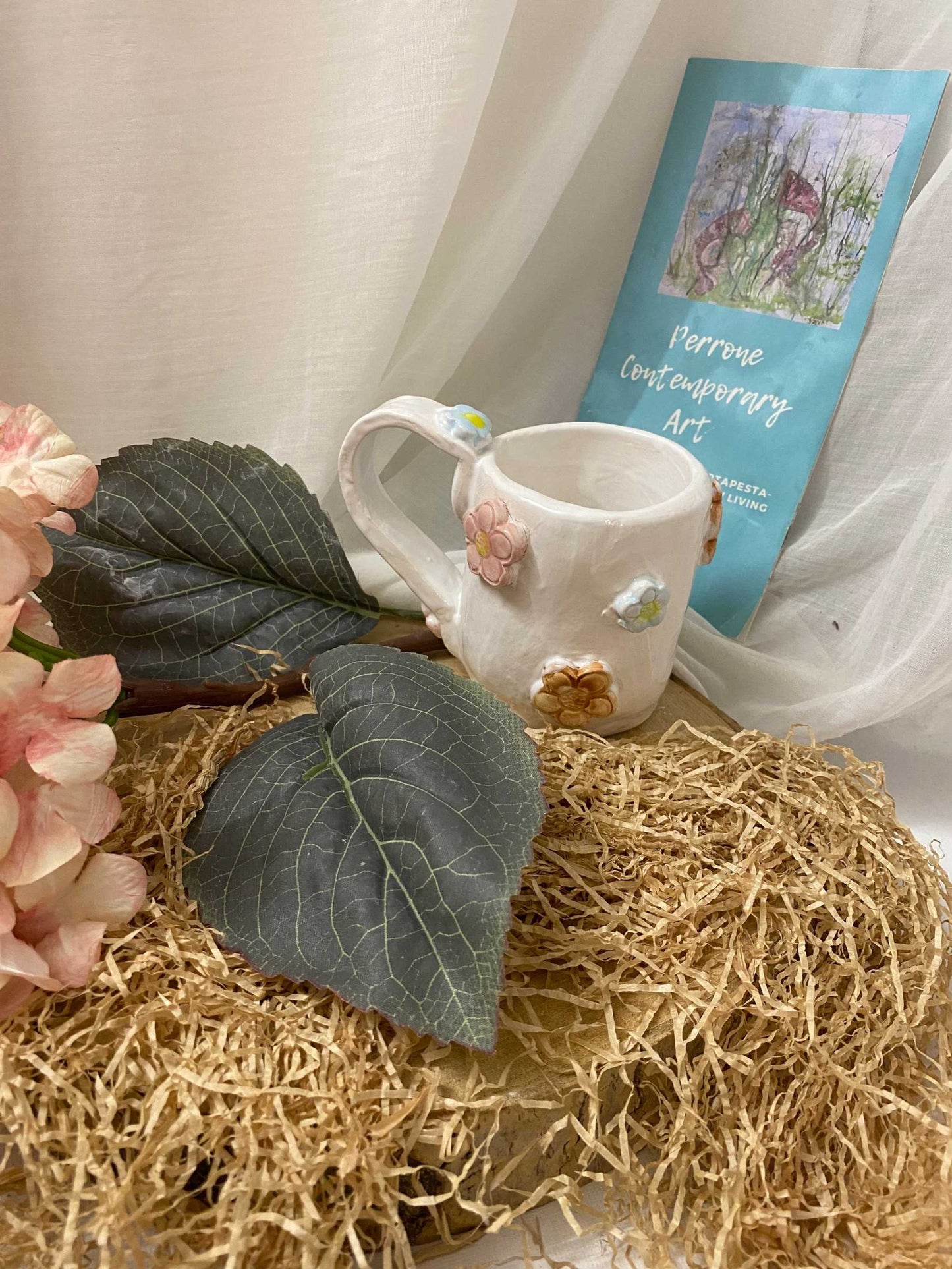 White ceramic Mug Collezione Primavera with floral decorations on a textured natural mat with leaves and a blue art book in the background