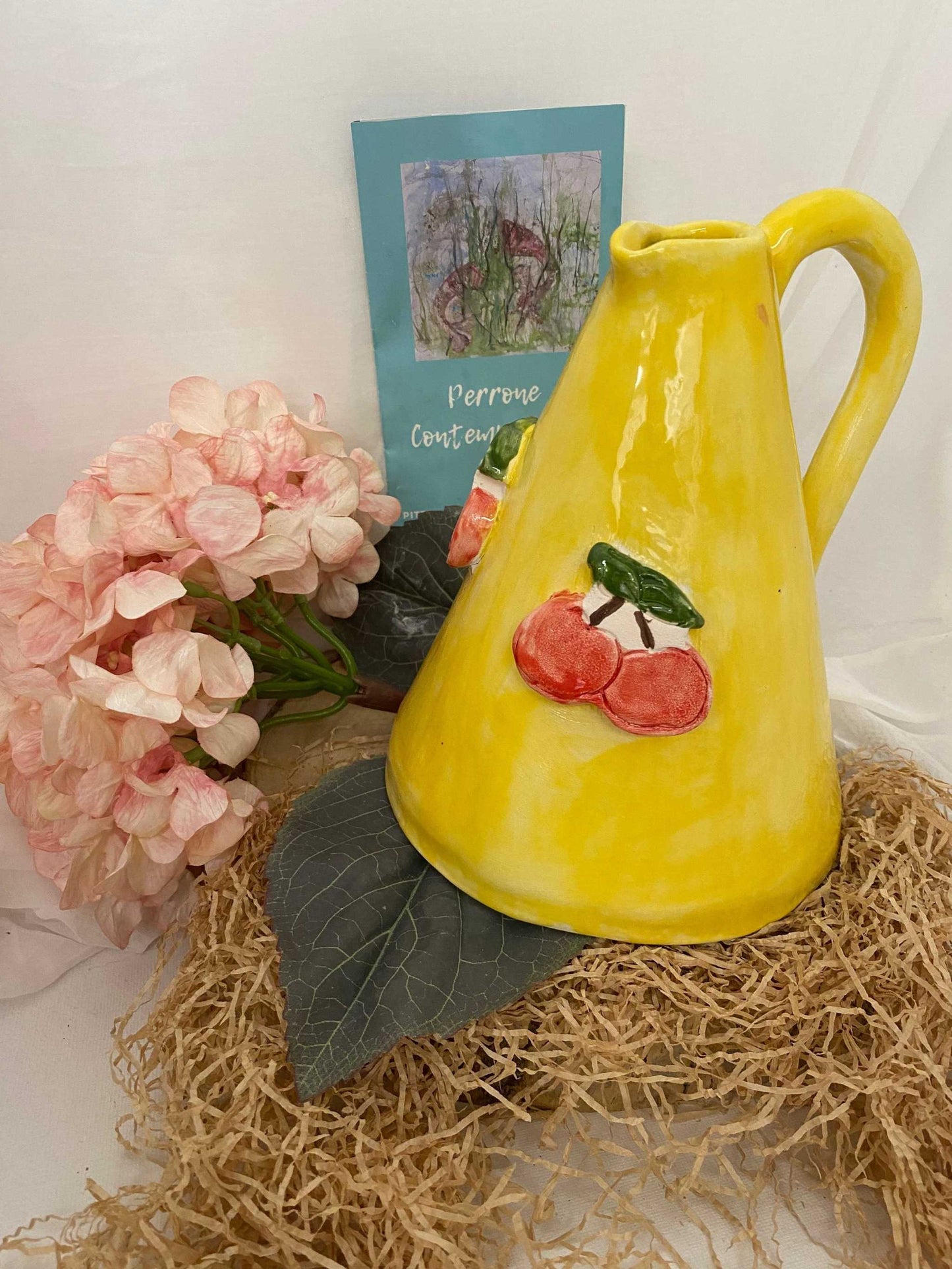Yellow ceramic olive oil dispenser with cherry decoration from Fruit Collection on woven mat with pink flowers and artistic card background