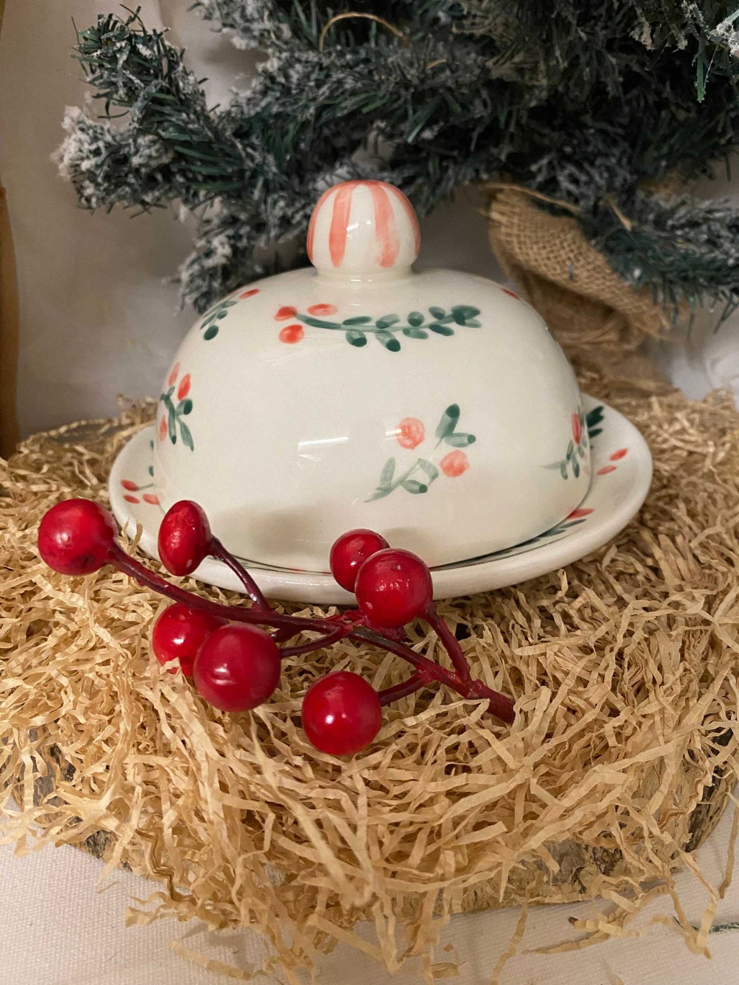 white ceramic butter dish with festive red and green floral design and knob handle on matching plate with decorative red berries and Christmas tree background