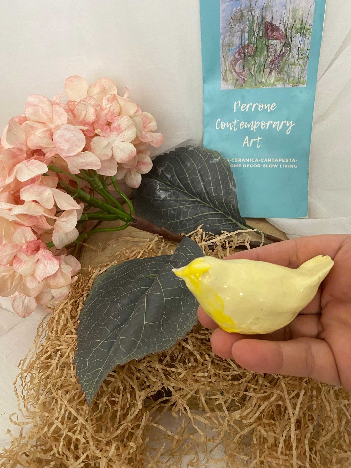Yellow ceramic bird from Primavera collection held in hand with decorative pink flowers and leaves in background